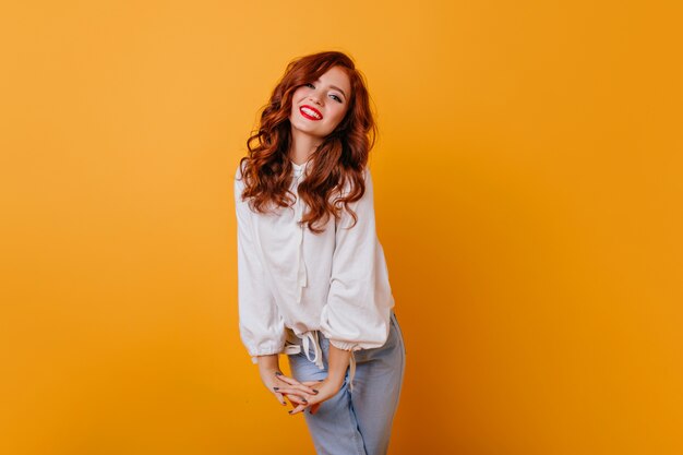 Jocund caucasin pani uśmiechnięta na pomarańczowej ścianie. Wewnętrzne zdjęcie wyrafinowanej kręconej kobiety w eleganckiej białej bluzce.