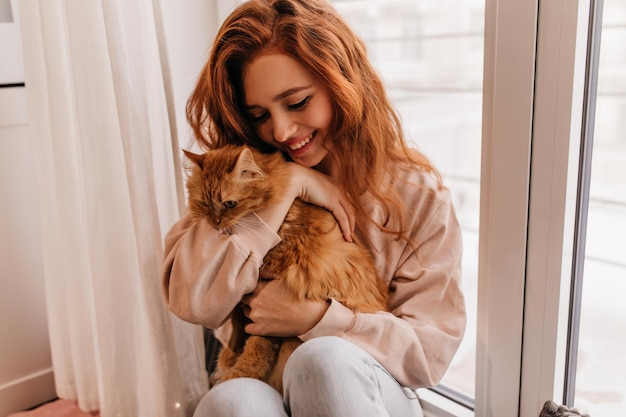 Jocund dama z falującymi włosami pozuje ze swoim zwierzakiem Wewnątrz portret europejskiej dziewczyny bawiącej się z kotem w domu