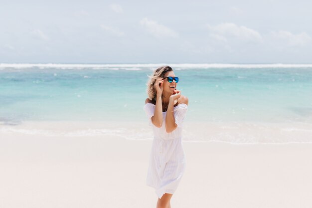 Jocund kobieta w romantyczny biały strój stojący na morzu. Roześmiana ekstatyczna kobieta w okularach przeciwsłonecznych spędzających letni dzień nad oceanem.