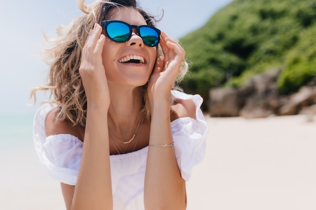 Jocund opalona dama w letnich ubraniach patrząc w niebo z radosnym uśmiechem. Zewnętrzne zdjęcie wesołej kobiety w eleganckich okularach przeciwsłonecznych korzystających z wypoczynku w ośrodku.