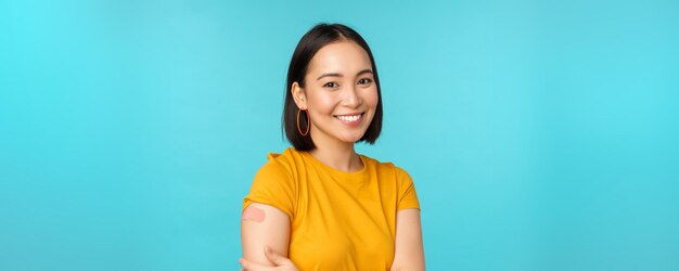 Kampania szczepionkowa od covid19 Młoda piękna zdrowa azjatycka kobieta pokazująca ramię z bandaidową koncepcją szczepienia stojącego na niebieskim tle