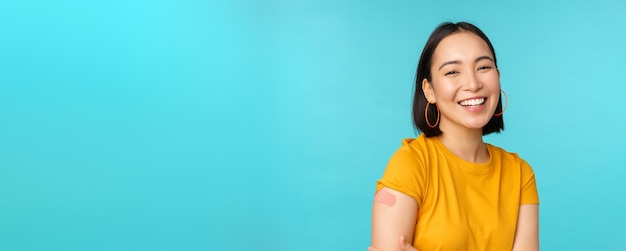 Kampania szczepionkowa od covid19 Szczęśliwa i zdrowa azjatycka dziewczyna śmiejąca się po szczepieniu z bandaża koronawirusa na ramieniu, ubrana w żółtą koszulkę na niebieskim tle
