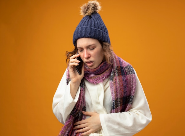 Bezpłatne zdjęcie kaszel młoda chora dziewczyna ubrana w białą szatę i czapkę zimową z szalikiem rozmawia przez telefon kładąc rękę na brzuchu odizolowaną na pomarańczowej ścianie