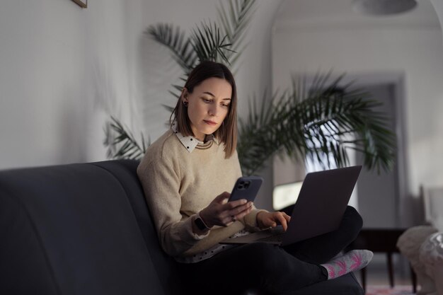 Kaukaska kobieta korzysta z laptopa siedząc na kanapie w domu wieczorem w salonie, pisząc na telefonie