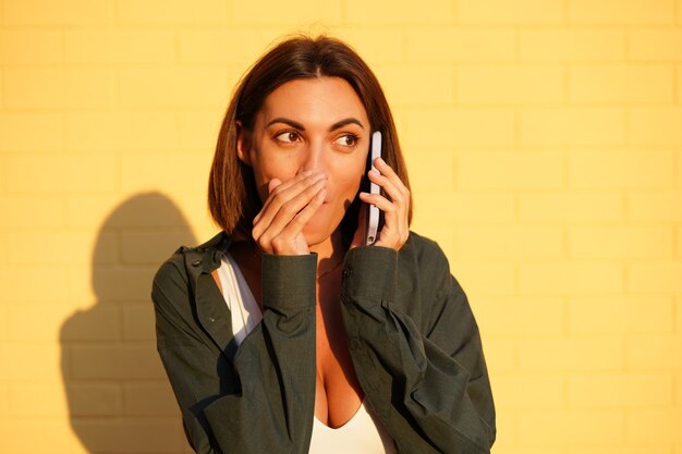 Kaukaska kobieta ubrana w koszulę o zachodzie słońca na żółtej ścianie z cegły na świeżym powietrzu pozytywne rozmowy na telefon komórkowy plotka mówiąca tajną osłonę ust ręką