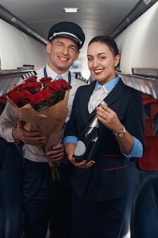 Kaukaski kapitan samolotu i hostessa uśmiechają się do kamery, trzymając butelkę szampana i bukiet róż