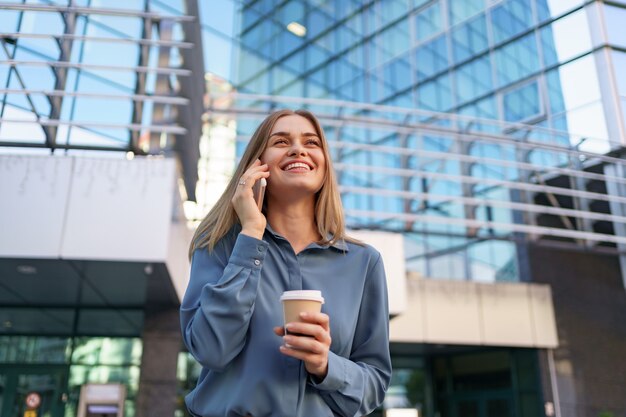 Kaukaski kobieta rozmawia przez telefon trzymając kawę, aby przejść. Europejka sukcesu, rozmawiająca przez telefon, stojąca na nowoczesnym biurowcu