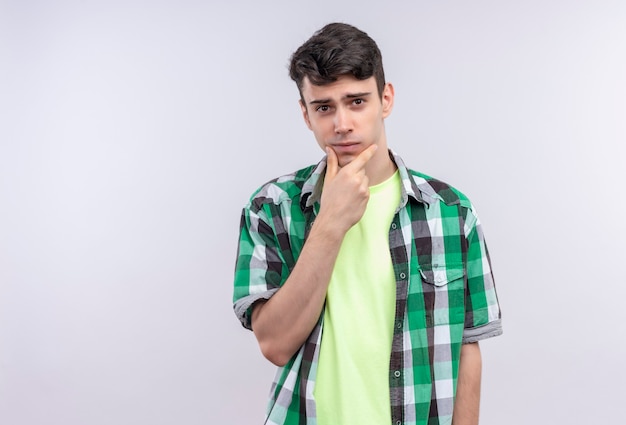 kaukaski młody człowiek ubrany w zieloną koszulę położył dłoń na brodzie na odosobnionej białej ścianie