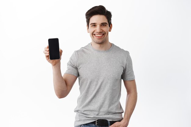 Kaukaski przystojny mężczyzna pokazuje ekran telefonu komórkowego, uśmiechając się i dając rekomendację aplikacji, stojąc w koszulce na białym tle