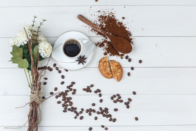 Kawa w filiżance z mieloną kawą, przyprawami, kwiatami, ziarnami kawy, ciasteczkami płasko leżała na drewnianym tle