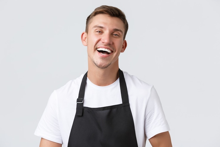 Bezpłatny plik Zdjęcie kawiarnie i restauracje, właściciele kawiarni i koncepcja sprzedaży detalicznej. zbliżenie wesoły przystojny sprzedawca w czarnym fartuchu zapraszający lub witający klientów w sklepie, śmiejący się szczęśliwy, białe tło