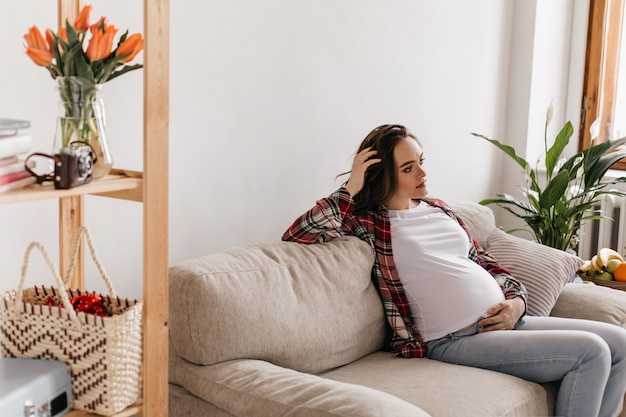 Kędzierzawa brunetka kobieta w ciąży w dżinsowych spodniach i koszuli w kratę spoczywa na miękkiej sofie