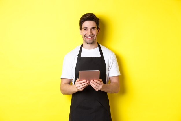 Kelner w czarnym fartuchu przyjmuje zamówienia, trzyma cyfrowy tablet i uśmiecha się przyjaźnie, stojąc na żółtym tle.