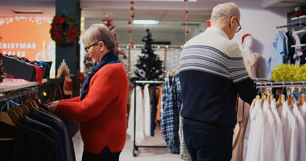 Klienci ośmiolatków przeglądający wieszaki na ubrania w świątecznym, ozdobnym sklepie z modą w okresie ferii zimowych. Starsza para jest szczęśliwa, gdy znalazła kolorowe marynarki na prezent na świątecznym spotkaniu rodzinnym