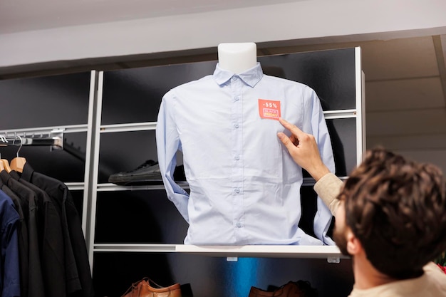 Bezpłatne zdjęcie klient płci męskiej sprawdza cenę koszuli, stojąc w sklepie odzieżowym i patrząc na czerwoną naklejkę z metką. klient liczący zniżki podczas zakupów w sklepie odzieżowym w czarny piątek