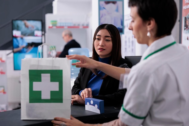 Klient Sklepu Aptecznego Kupuje Witaminy I Płaci Przy Kasie W Aptece. Asystent Farmaceutyczny Sprzedający Suplement Młodemu Azjatyckiemu Klientowi I Pakujący Pakiet Multiwitaminowy