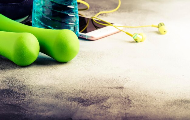 Kobieca koncepcja fitness jasnozielone hantle izotoniczny niebieski smartfon z wodą ze słuchawkami i butami do biegania na szarej betonowej podłodze selektywne skupienie