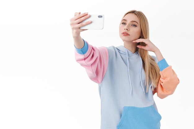 Kobieca, ładna blondynka o zmysłowym wyrazie twarzy, delikatnie dotykająca twarzy podczas robienia selfie na telefonie komórkowym