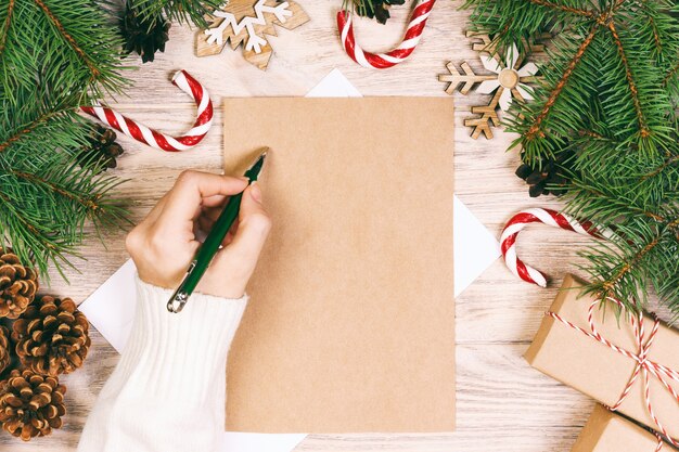 Kobiece ręce pisząc list do świętego mikołaja. prezenty świąteczne z dekoracją świąteczną na drewnianym tle. widok z góry. stonowany.