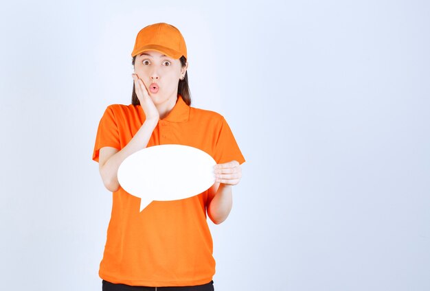 Kobieta agentka w pomarańczowym dresscode trzyma owalną tablicę informacyjną i wygląda na zaskoczoną i przerażoną