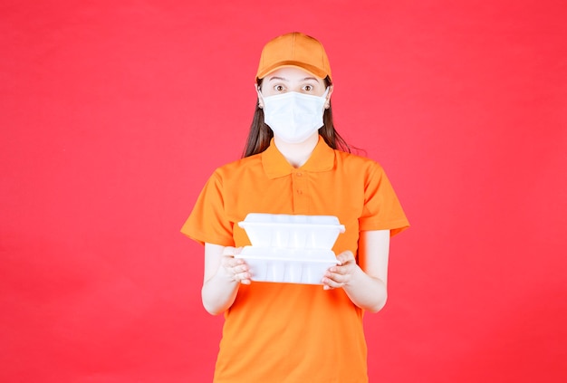 Kobieta agentka w pomarańczowym ubiorze i masce, trzymająca dwa opakowania z jedzeniem na wynos