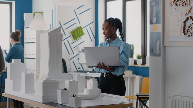 Kobieta architekt posiadający laptopa do analizy modelu budynku w biurze architektonicznym. Inżynier pracujący z komputerem i makietą przy projektowaniu konstrukcji budowlanych dla projektu urbanistycznego.