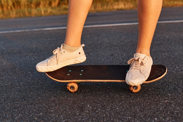 Kobieta bez twarzy w białych butach jeżdżąca na longboardzie po asfalcie, nieznana osoba jeździ samotnie na deskorolce, dziewczyny nogi na deskorolce.