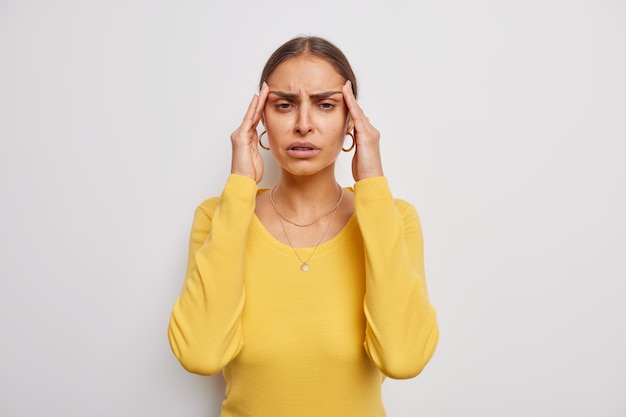 kobieta cierpi na ból głowy trzyma ręce na skroniach sfrustrowana porażką grymasy z bólu potrzebuje środków przeciwbólowych nosi swobodny żółty sweter na białym tle