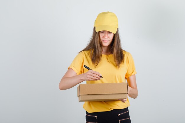 Kobieta dostawy w t-shirt, spodnie, czapka pisząca na tekturowym pudełku i wyglądająca na zajętą