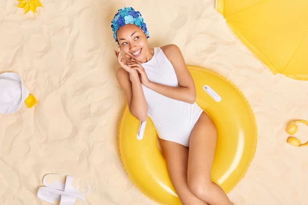 Kobieta Dotyka Twarzy Delikatnie Odwraca Wzrok Szczęśliwie Nosi Niebieskie Nakrycie Głowy Biały Strój Kąpielowy Siedzi Na żółtym Napompowany Pływak Ma Marzycielski Wyraz Relaksuje Się Na Plaży