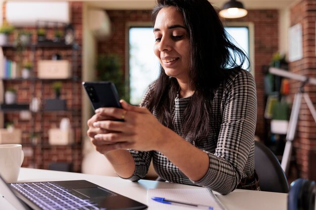 Kobieta freelancer pracująca w aplikacji na smartfona i przy użyciu laptopa do pracy zdalnej na biurku w domu. Przeglądanie internetu i mediów społecznościowych na telefonie komórkowym do wiadomości tekstowych, przerwa na badania online.