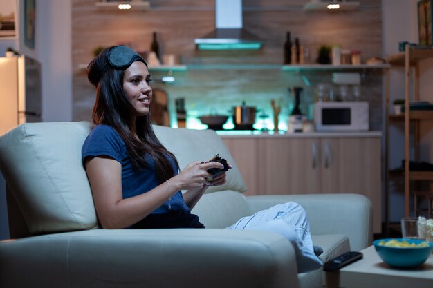 Kobieta grająca w domu, siedząca na kanapie, grająca w gry wideo późno w nocy, nosząca maskę na oczy na czole