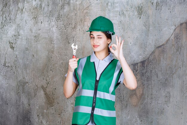 Kobieta inżynier w zielonym kasku trzymająca metalowy klucz do naprawy i pokazująca znak przyjemności