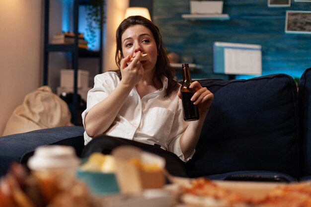 Kobieta je kawałek gorącej pizzy dostawy siedzi na kanapie trzymając butelkę piwa patrząc na telewizor w salonie. Osoba po pracy korzystająca z telewizji na wynos kolacja przy stole z fast foodami na wynos.