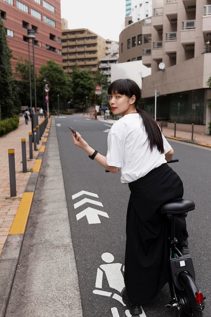 Kobieta jedzie na rowerze elektrycznym po mieście i trzyma smartfon