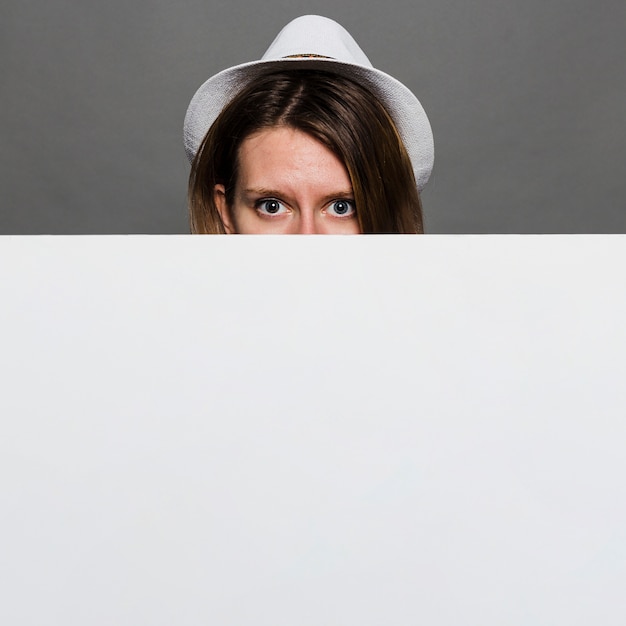 Kobieta jest ubranym białego kapeluszu zerkanie przez białej pustej karty przeciw szarej ścianie