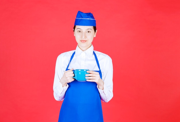 Bezpłatne zdjęcie kobieta kelnerka w mundurze stojąc i trzymając niebieski kubek na czerwonej ścianie.