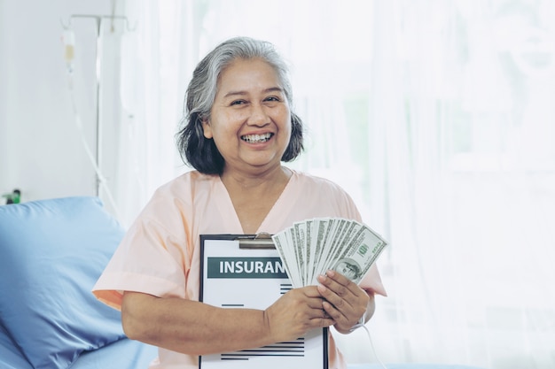 Kobieta, która doznała uszczerbku na zdrowiu osób starszych na łóżku pacjenta w szpitalu, trzymając nas banknotów dolarowych, czuje się szczęśliwa z otrzymywania pieniędzy ubezpieczeniowych od firm ubezpieczeniowych