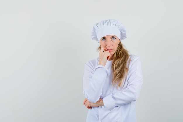 Kobieta kucharz stojąc w myśleniu poza w białym mundurze i patrząc bezradnie.
