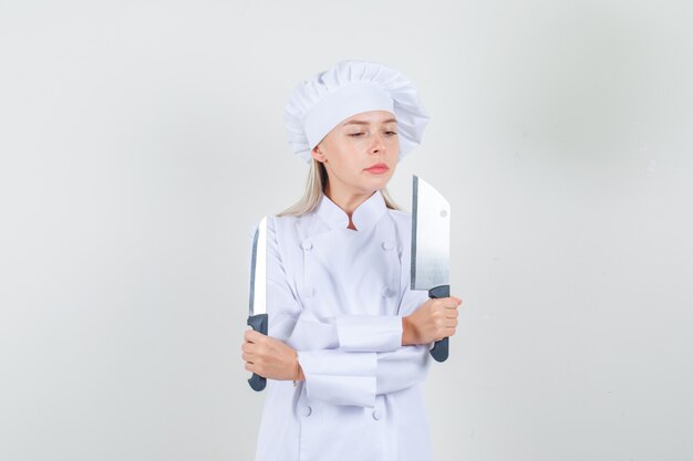 Kobieta kucharz trzyma nóż i tasak w białym mundurze i wygląda poważnie