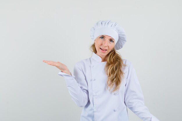 Kobieta kucharz w białym mundurze, rozkładająca dłoń na bok i wyglądająca wesoło