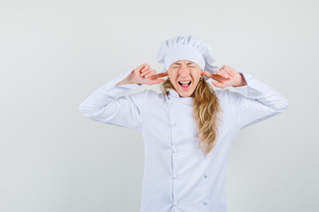 Bezpłatne zdjęcie kobieta kucharz zatyka uszy palcami w białym mundurze i wygląda na zirytowaną