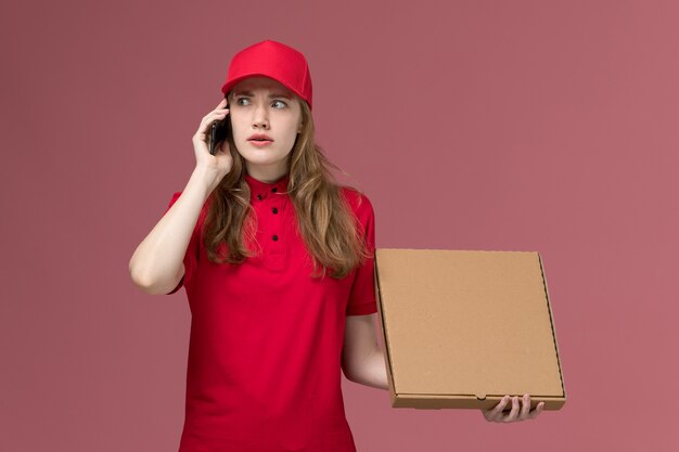 kobieta kurier w czerwonym mundurze rozmawia przez telefon trzymając pudełko z jedzeniem na różowym, jednolita praca pracownika dostawy usług