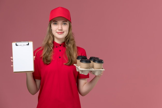 kobieta kurier w czerwonym mundurze uśmiechnięty trzymając notatnik i filiżanki kawy na różowym, jednolitej pracy pracownika dostawy usług
