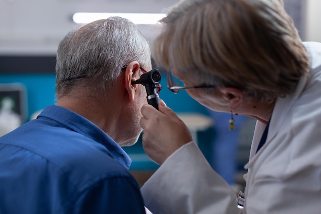 Kobieta lekarz posiadający otoskop do konsultacji ucha dla starego człowieka na wizytę lekarską. Otolog za pomocą przyrządu otologicznego do wykonania badania i udzielenia porady medycznej choremu pacjentowi.