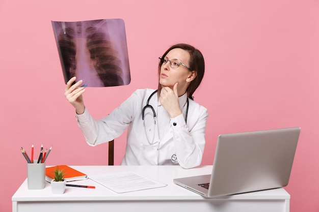 Kobieta lekarz siedzi przy biurku w pracy na komputerze z dokumentem medycznym trzymać rtg w szpitalu na białym tle na pastelowym różowym tle ściany. kobieta w medycznym stetoskopie toga okularów. koncepcja medycyny opieki zdrowotnej.