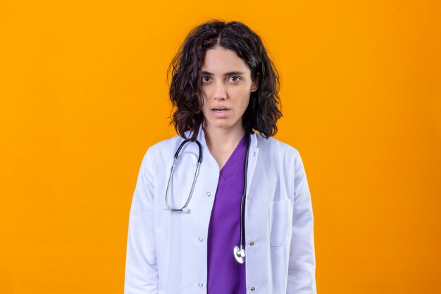 kobieta lekarz ubrany w biały fartuch ze stetoskopem bez uśmiechu stojącego na odizolowanych pomarańczy