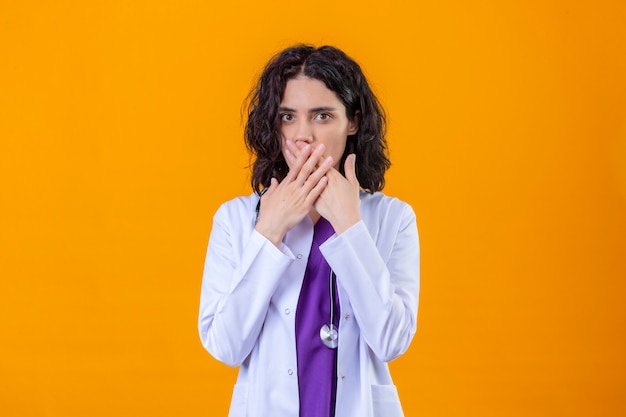 kobieta lekarz ubrany w biały fartuch ze stetoskopem, patrząc zaskoczony ustami stożkowymi rękami stojącymi na odosobnionym pomarańczy