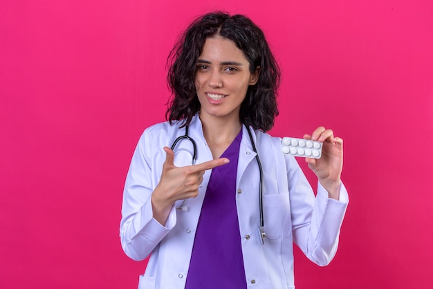 kobieta lekarz ubrany w biały fartuch ze stetoskopem wskazującym palcem na blister z pigułkami z uśmiechem na twarzy na odosobnionym różu