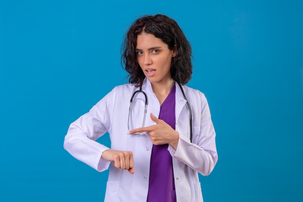 Kobieta Lekarz W Białym Fartuchu Ze Stetoskopem Wskazującym Na Jej Dłoń Przypominająca O Czasie Ze Sceptycznym Wyrazem Twarzy Stojącej Na Odosobnionym Błękicie